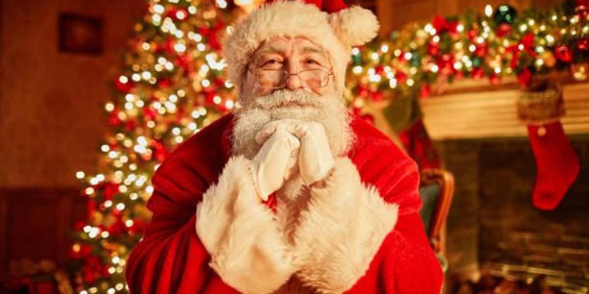 Retrato de frente del tradicional Papá Noel sonriendo a la cámara apoyando la cabeza en los brazos con el árbol de navidad de fondo.