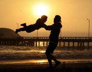 Imagen de un padre jugando con su hijo, alzándolo con sus brazos, en una playa.