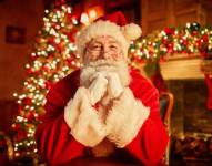 Retrato de frente del tradicional Papá Noel sonriendo a la cámara apoyando la cabeza en los brazos con el árbol de navidad de fondo.