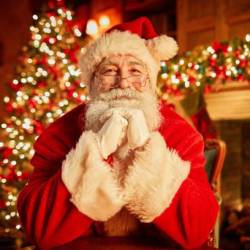 Retrato de frente del tradicional Papá Noel sonriendo a la cámara apoyando la cabeza en los brazos con el árbol de navidad de fondo.