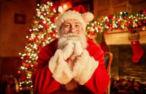 Retrato de frente del tradicional Papá Noel sonriendo a la cámara apoyando la cabeza en los brazos con el árbol de navidad de fondo.