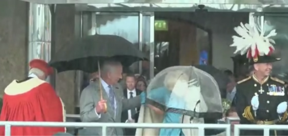 Un asistente real ayuda a la reina Camila a colocarse su abrigo, en una tormenta registrada en Jersey, donde disfrutaban de un desfile, después del pedido del rey Carlos III.
