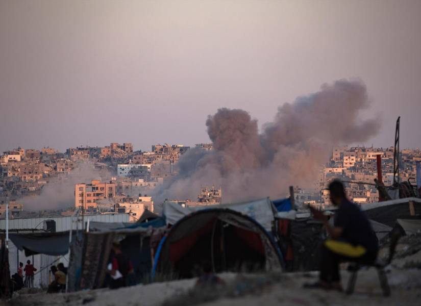 El humo se eleva tras un ataque aéreo israelí, mientras los palestinos desplazados internos se sientan junto a sus tiendas de campaña en el campamento.